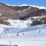 Skiers on a sunny winter slope in Campitello Matese, with snow-covered mountains and chairlifts in the background.