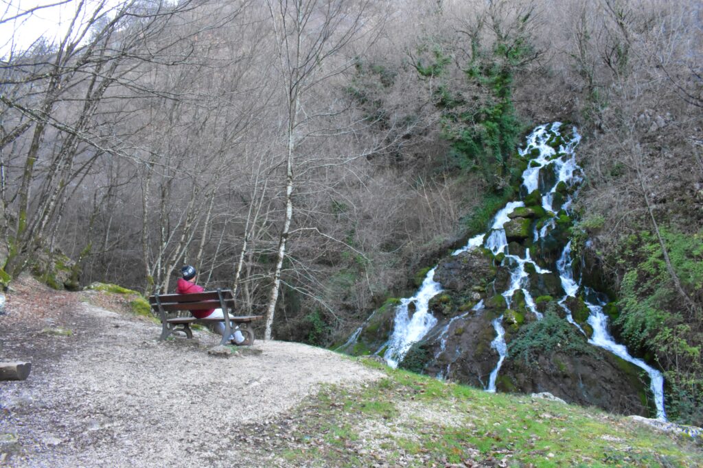 Person sitting on a bench overlooking the Cascate Rio waterfall in Molise, surrounded by bare winter trees and moss-covered rocks.