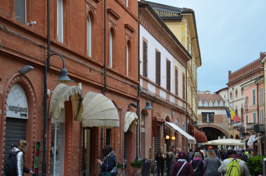 A lively street scene in Ravenna with people walking past red-brick and pastel-colored buildings lined with shops and cafes. Some people are chatting or taking photos, and flags are visible in the background near an arched gateway.