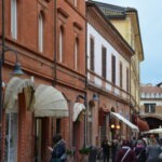 A lively street scene in Ravenna with people walking past red-brick and pastel-colored buildings lined with shops and cafes. Some people are chatting or taking photos, and flags are visible in the background near an arched gateway.