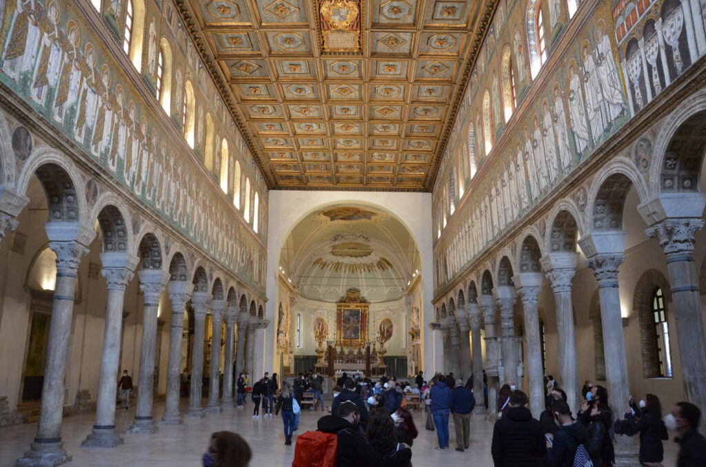 „Wnętrze bazyliki Sant’Apollinare Nuovo w Rawennie – długa nawa z kolumnadą i słynnymi bizantyjskimi mozaikami ukazującymi procesję świętych.