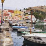 Colorful waterfront of Procida Island, Italy, with pastel-colored houses rising up the hillside, small boats moored along the harbor, and a historic fortress overlooking the town.