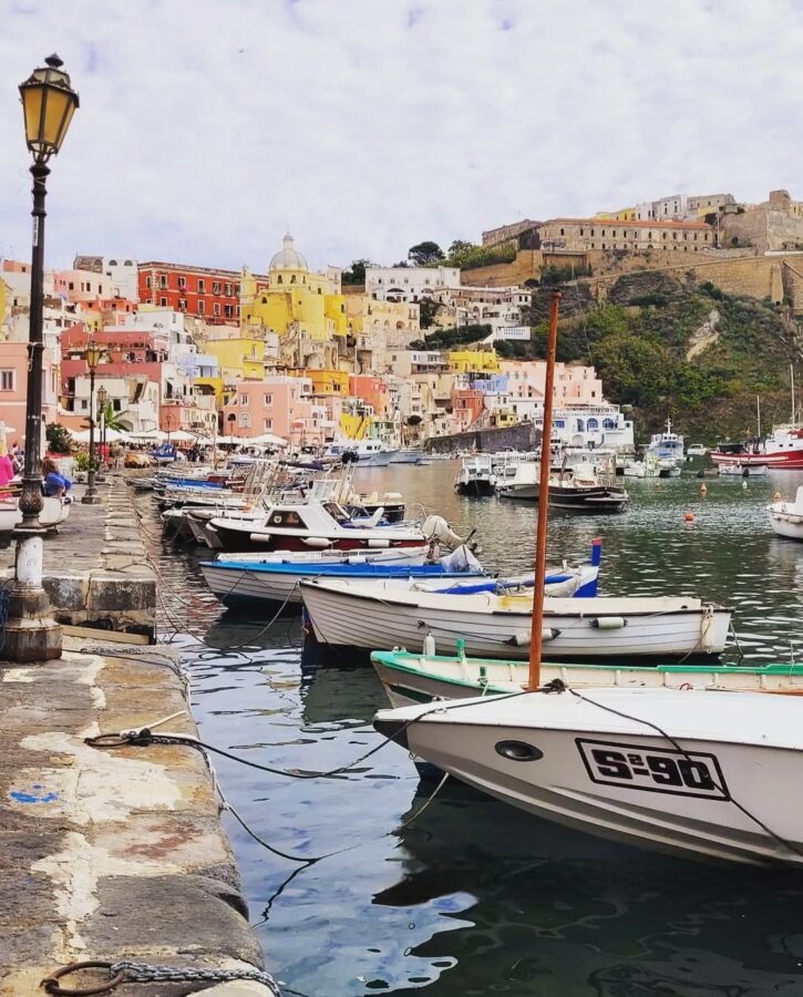 Colorful waterfront of Procida Island, Italy, with pastel-colored houses rising up the hillside, small boats moored along the harbor, and a historic fortress overlooking the town.
