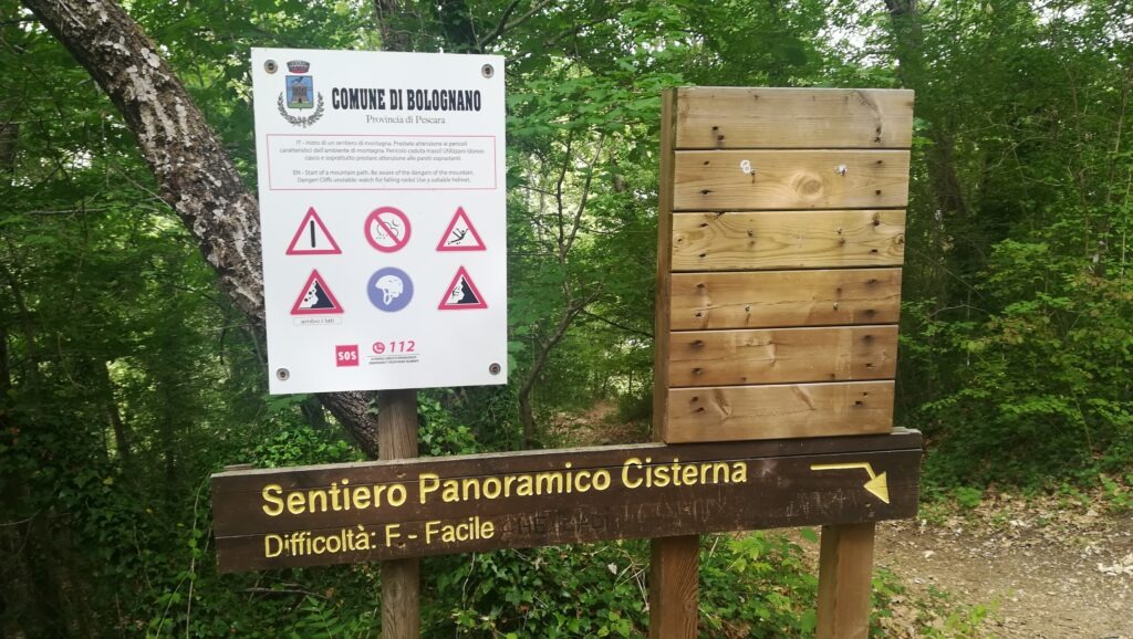 Informational signs at the start of the Sentiero Panoramico Cisterna in Bolognano, Abruzzo. A white warning board from the Comune di Bolognano displays safety icons for falling rocks and unstable cliffs, while a wooden sign below indicates the trail is "Difficoltà: F - Facile" (Easy) with a yellow arrow pointing toward a forest path.