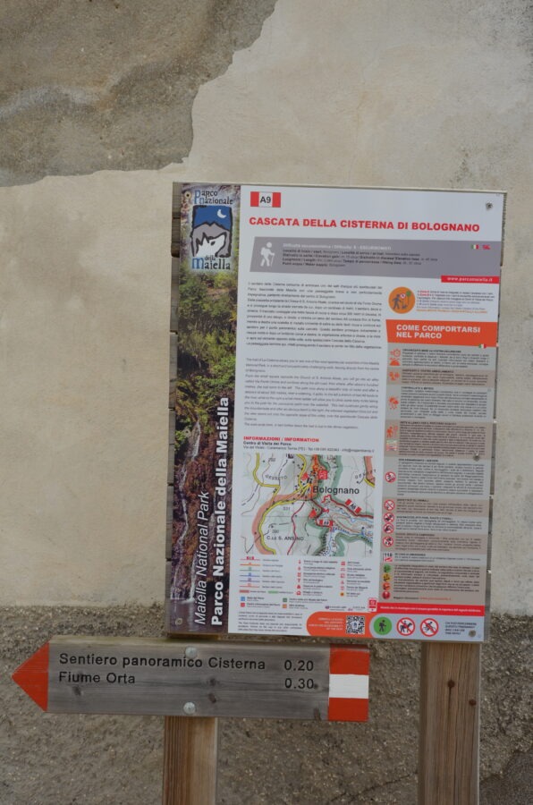 Wooden trail sign and information board for Cascata della Cisterna di Bolognano in Maiella National Park. The sign points toward "Sentiero panoramico Cisterna" (20 minutes) and "Fiume Orta" (30 minutes), with a detailed map and park regulations displayed on the board above.