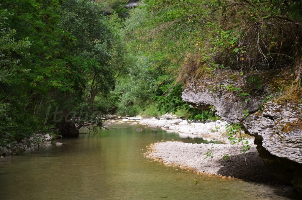 A scenic view of the calm Orta River flowing through a lush green canyon in Abruzzo. The water is clear and surrounded by dense trees, white river stones on the banks, and a prominent overhanging limestone rock formation on the right side.