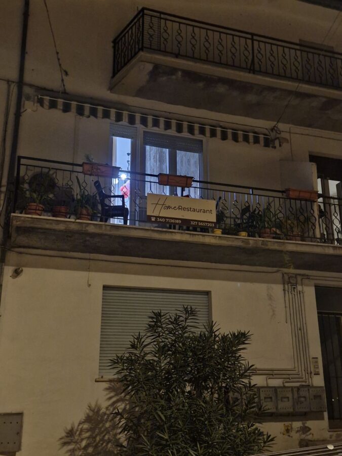 Evening view of a small apartment balcony in Palombaro, Italy, with a “Home Restaurant” sign, potted plants on the railing, and light coming from the open doorway, hinting at the cozy restaurant inside.