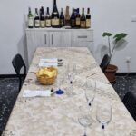 Dining table set with glasses, napkins, and a basket of bread at Home Restaurant da Elisa in Palombaro, Italy, with a cabinet displaying assorted wine bottles in the background.