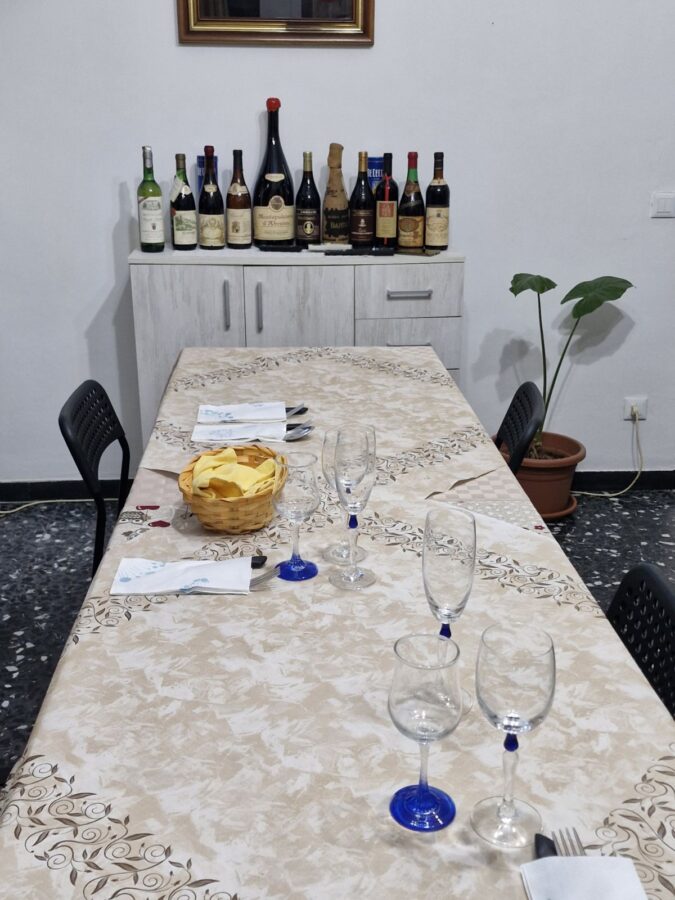 Dining table set with glasses, napkins, and a basket of bread at Home Restaurant da Elisa in Palombaro, Italy, with a cabinet displaying assorted wine bottles in the background.