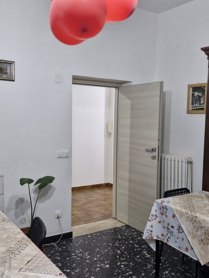 Interior of Home Restaurant da Elisa in Palombaro, Italy, showing simple home-style decor with red balloons hanging from the ceiling, a doorway, and tables covered with patterned tablecloths.