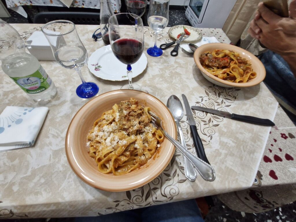 Table set for dinner at Home Restaurant da Elisa in Palombaro, Italy, with bowls of fresh pasta topped with sauce and cheese, glasses of red wine, and simple home-style table settings.