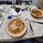 Table set for dinner at Home Restaurant da Elisa in Palombaro, Italy, with bowls of fresh pasta topped with sauce and cheese, glasses of red wine, and simple home-style table settings.