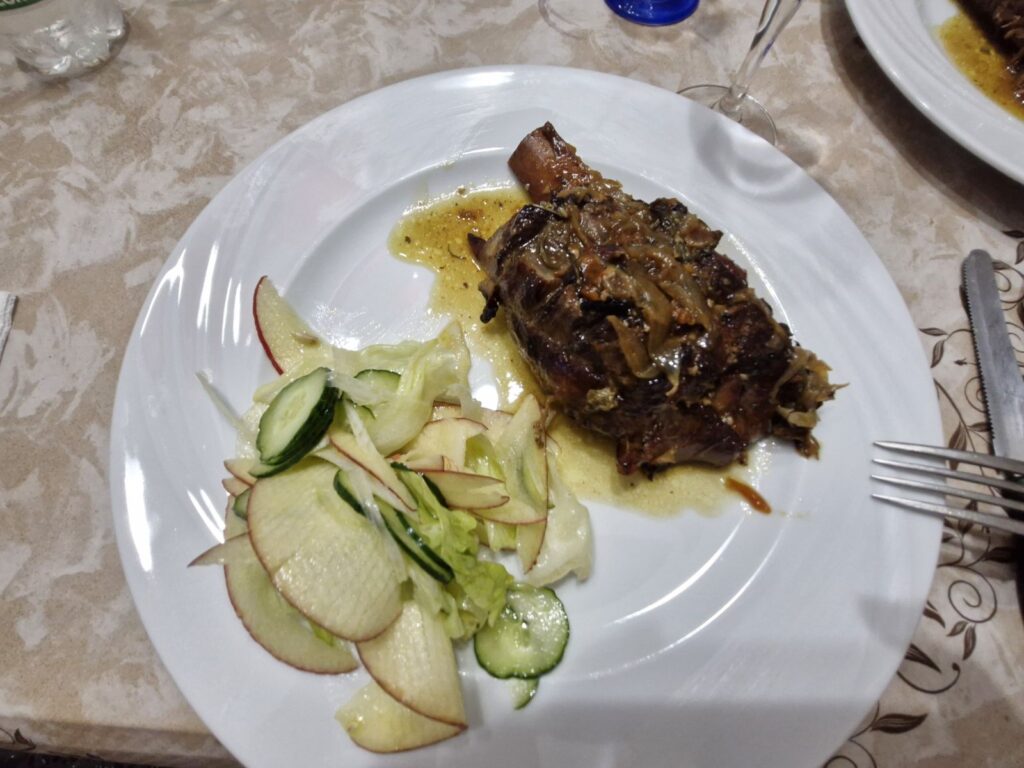 Plate of roasted pork knuckle in sauce served with a fresh side salad of sliced apples, cucumbers, and lettuce on a white tablecloth at Home Restaurant da Elisa in Palombaro, Italy.