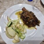 Plate of roasted pork knuckle in sauce served with a fresh side salad of sliced apples, cucumbers, and lettuce on a white tablecloth at Home Restaurant da Elisa in Palombaro, Italy.