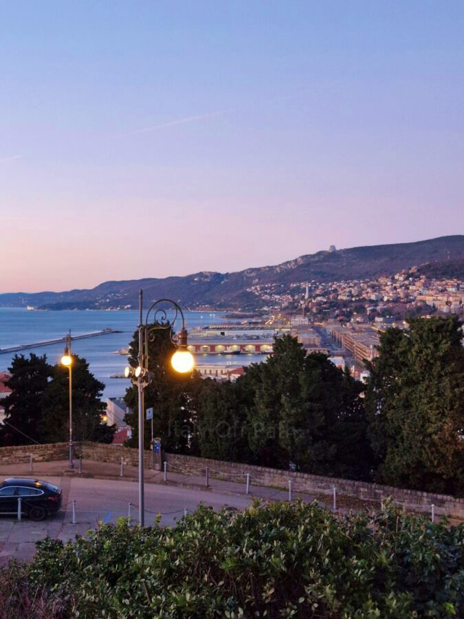 Panorami z góry na Triest o zmierzchu. Na pierwszym planie widać zapaloną, ozdobną latarnię uliczną oraz zielone krzewy i drzewa. W tle rozciąga się zatoka z portem, zabudowania miasta na wzgórzach oraz pastelowe, fioletowo-różowe niebo tuż po zachodzie słońca.