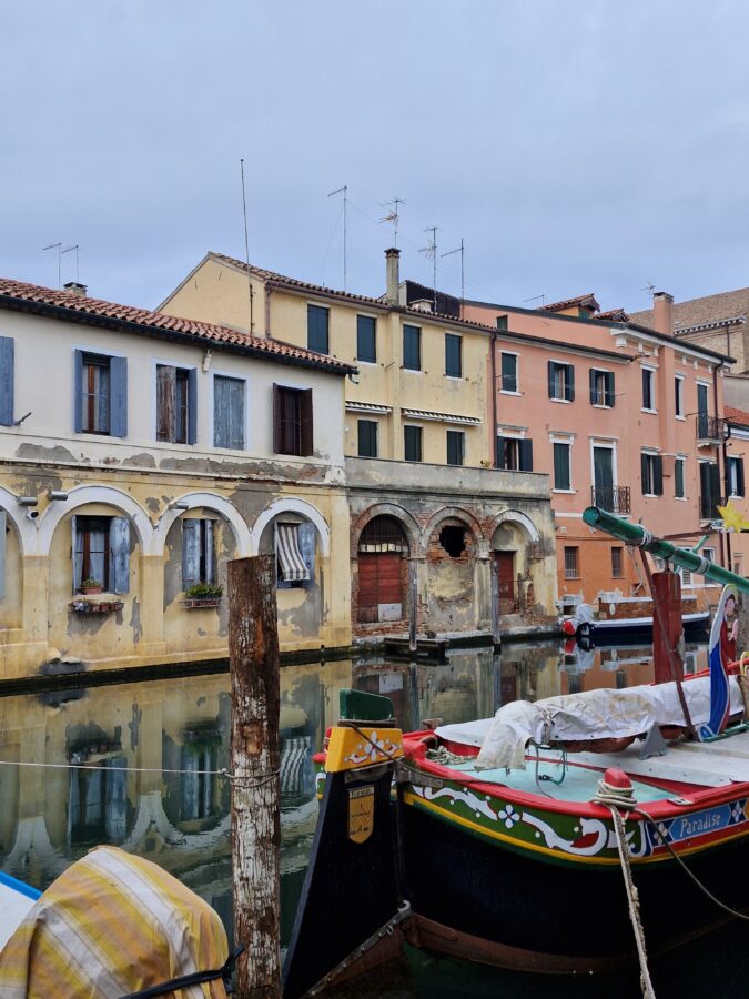 A traditional, brightly painted wooden boat named "Paradiso" moored in a canal in Chioggia, Italy. In the background, weathered multi-story buildings with arched loggias, peeling yellow and peach plaster, and dark window shutters are reflected in the calm water under a grey sky.