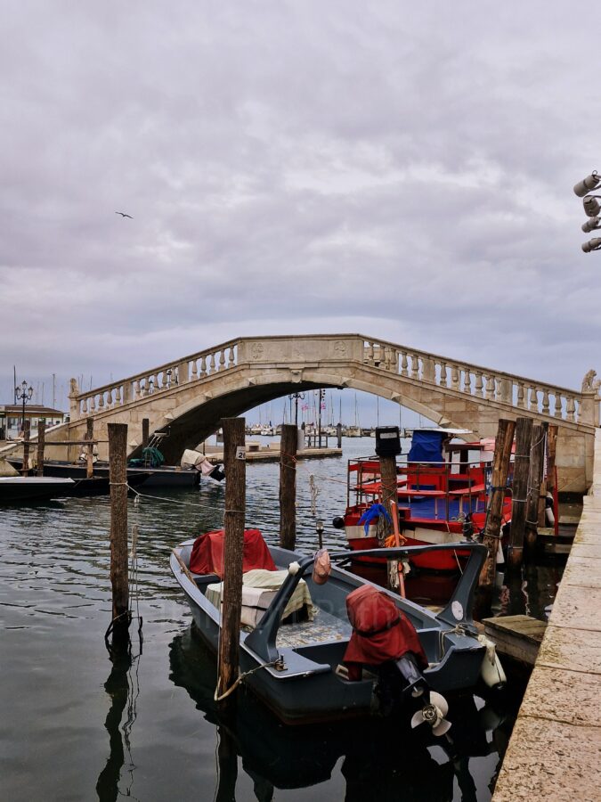 A stone arch bridge with classic balustrade railings spanning over a canal entry in Chioggia, Italy. Several small motorboats and a larger red-and-white vessel are moored to wooden poles in the foreground water. Through the archway, a harbor with sailboat masts is visible under a heavy, overcast grey sky.