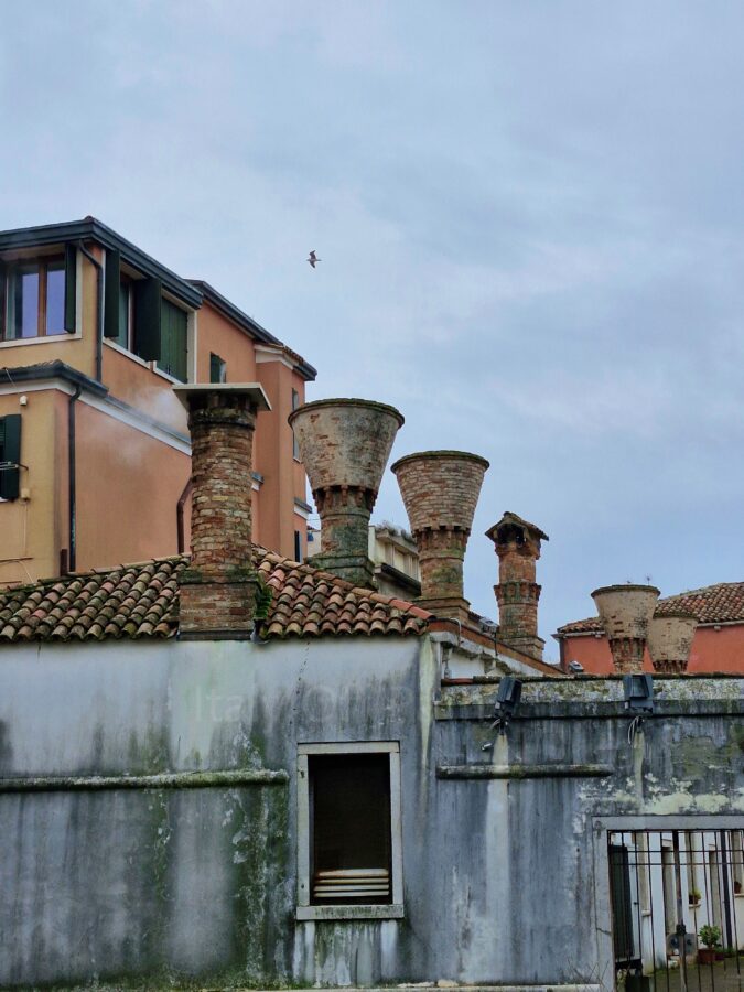 Visitong Chioggia. A close-up view of traditional Venetian-style chimneys on roofs in Chioggia, Italy. The foreground features weathered brick chimneys, including two distinct inverted cone-shaped (bell-shaped) designs, rising from a terracotta tiled roof on a stained, grey plaster building. A single bird flies in the overcast grey sky above, with multi-story peach and orange residential buildings visible in the background.