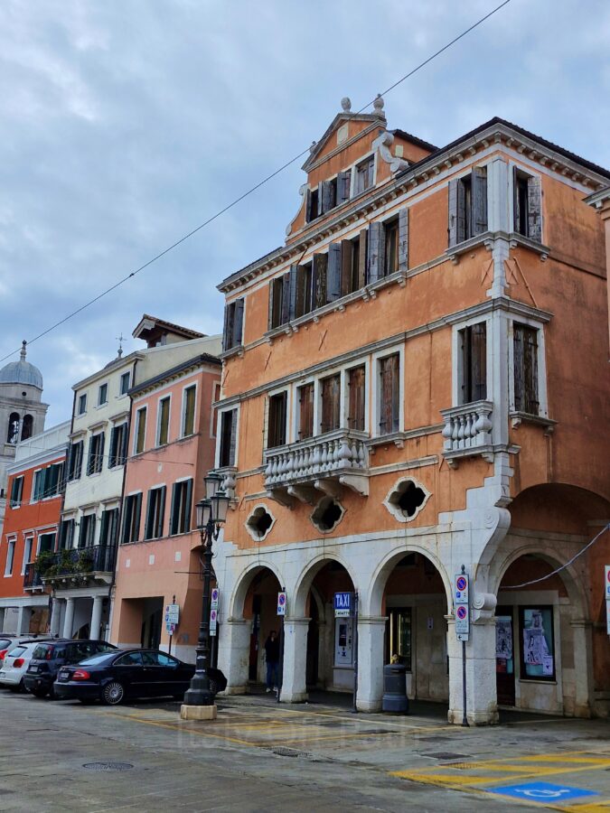A row of multi-story, colorful buildings in Chioggia, Italy, featuring a prominent terracotta-orange palazzo with stone arches, stone balconies, and dark wooden window shutters. In the background, other houses in shades of pink, yellow, and red line the street alongside parked cars under an overcast sky, with a white church dome visible in the distance.
