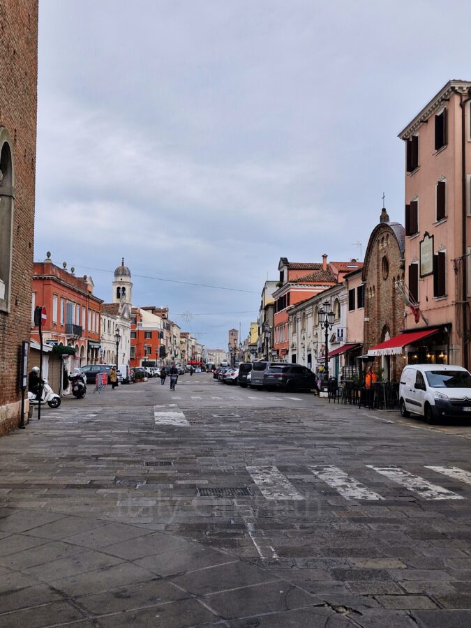 A wide, stone-paved street stretching into the distance under an overcast sky in Chioggia, Italy. The street is lined with multi-story buildings in shades of pink, terracotta, and white, including a brick church facade with a red awning and a hotel on the right. Parked cars, a white van, a scooter, and a few pedestrians are scattered along the sides of the street, with a church tower visible further down on the left.