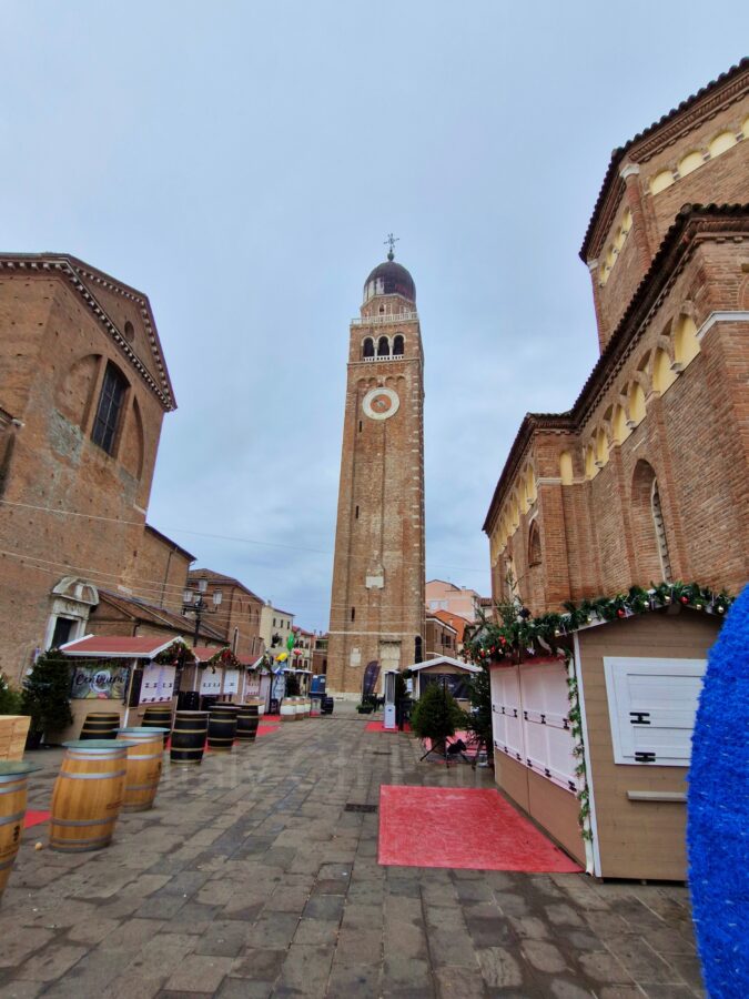 A tall, historic brick clock tower with a dark dome stands centrally at the end of a stone-paved square in Chioggia, Italy. Large brick buildings frame the left and right sides of the square, where wooden market stalls decorated with festive garlands, red carpets, and wooden barrels are set up under a cloudy sky.