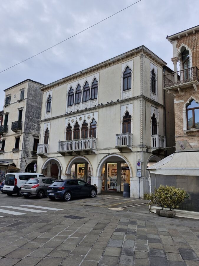A three-story Gothic-style building in Chioggia, Italy, featuring a light cream facade, ground-floor stone arches housing a bookstore, and ornate Venetian pointed-arch windows with stone balconies. Pigeons are lined up along the building's mid-level ledge, and several cars are parked on the paved street out front under a cloudy sky.