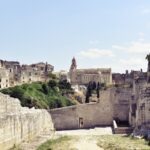 A view from a stone path in the Gravina di Puglia canyon looking toward the historic cathedral and old town buildings under a clear sky.