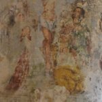 A weathered, ancient religious fresco of the crucifixion painted directly onto the limestone wall of a cave church in Gravina di Puglia