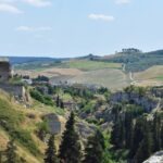 A vertical shot looking down into the lush green canyon of Gravina di Puglia with limestone cliffs and cypress trees.