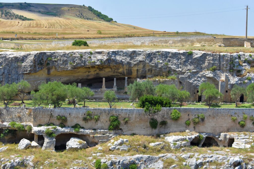 View of the Sette Camere rock complex in Gravina di Puglia, featuring caves and chambers carved into the pale limestone ravine slope, with visible stone columns supporting a rock overhang, surrounded by olive trees and sparse greenery under a clear sky.