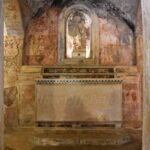 " A stone altar inside a cave church in Gravina di Puglia with faded frescoes and a religious statue in a niche.