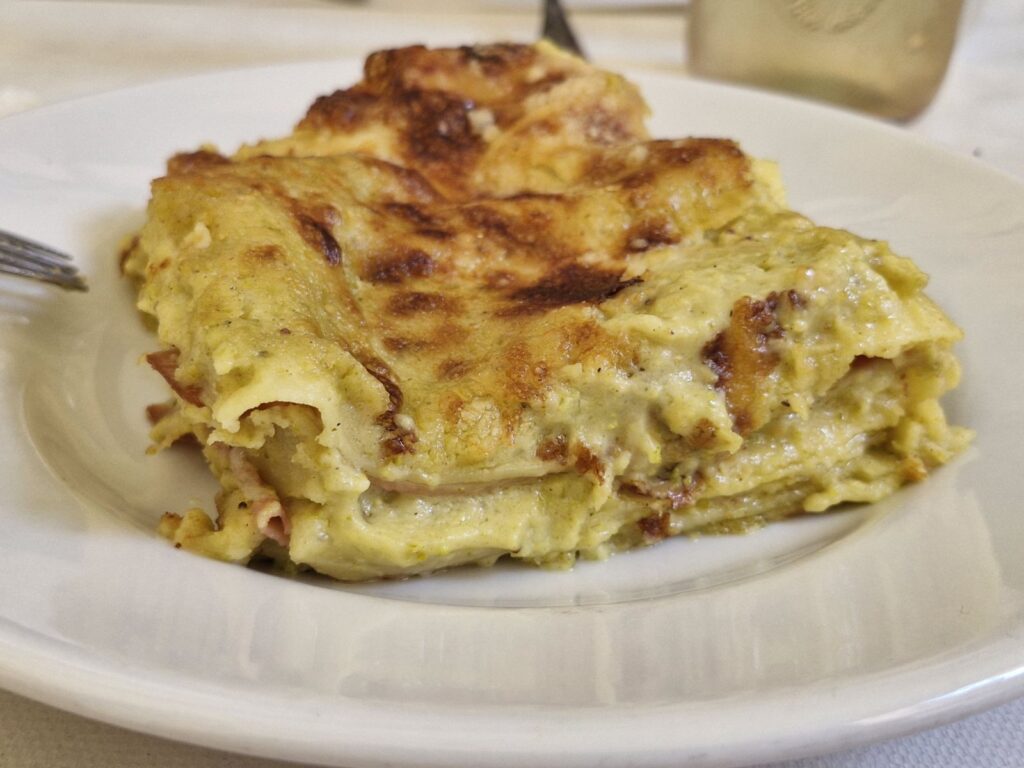 A close-up shot of a thick square of baked pistachio and mortadella lasagne served on a white plate. The layers show a creamy green pistachio sauce and thin slices of mortadella, topped with a golden-brown, crispy cheese crust.