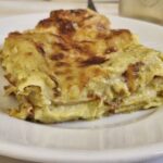 Porcja zapiekanej lasagne na białym talerzu – warstwy makaronu z kremowym sosem, serem i kawałkami mortadeli, lekko przypieczona na wierzchu.