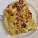 A slice of baked lasagna on a white plate, topped with a golden, slightly browned cheese crust, with visible creamy layers and a fork placed beside it.