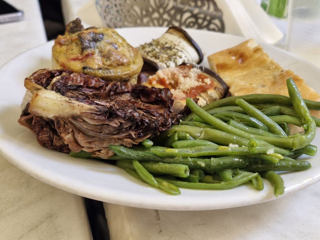 Talerz z różnymi daniami: zielona fasolka, pieczone warzywa (bakłażan, karczoch), kawałek zapiekanki warzywnej oraz kawałek focaccii, podane w stylu stołówkowym.