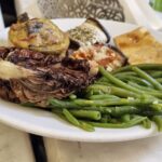 Talerz z różnymi daniami: zielona fasolka, pieczone warzywa (bakłażan, karczoch), kawałek zapiekanki warzywnej oraz kawałek focaccii, podane w stylu stołówkowym.