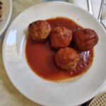 Plate of four meatballs in rich tomato sauce, served on a white dish, with part of a vegetable side visible in the background.
