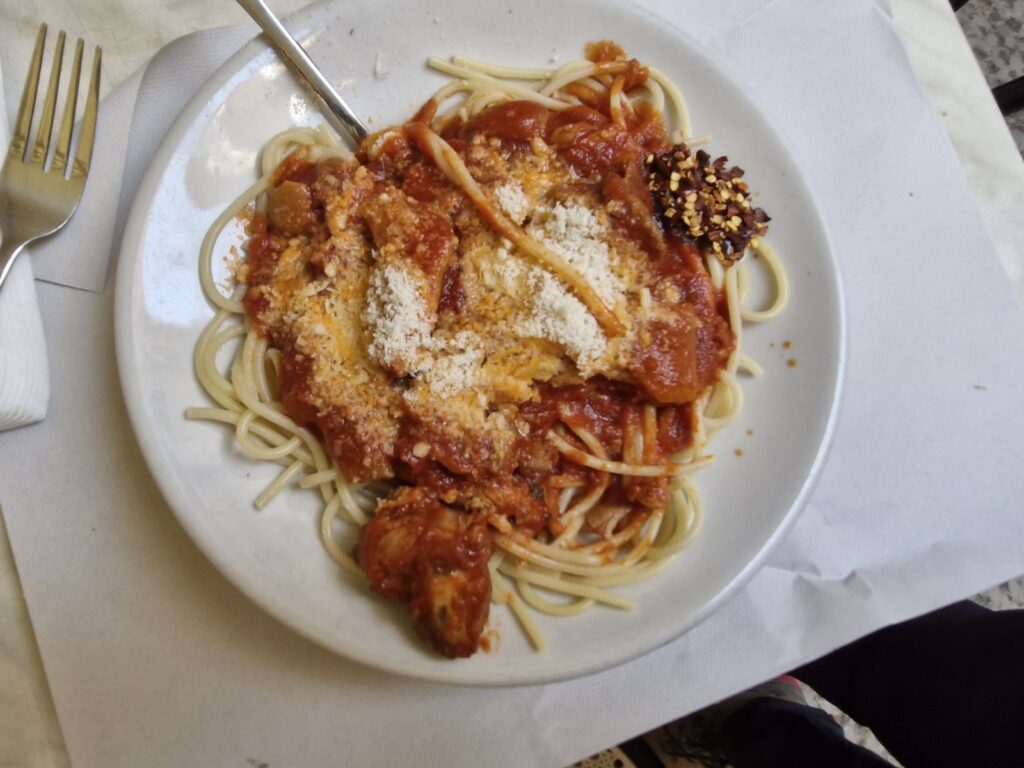 A plate of Sicilian pasta served at Basile Focacceria del Massimo, featuring long pasta topped with a rich tomato sauce, pieces of meat, pork rind, and a generous dusting of grated cheese. A small pile of dried chili flakes sits on the side of the white plate, which is placed on a simple paper table mat.