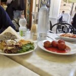 Where locals eat in Palermo. An outdoor restaurant table in Palermo featuring plates of traditional Sicilian food, including large meatballs in tomato sauce, a pasta dish, and a plate with green beans, grilled vegetables, and a slice of focaccia. Two bottles of water and a glass carafe sit on the marble table, with the entrance to Focacceria del Massimo visible in the background.