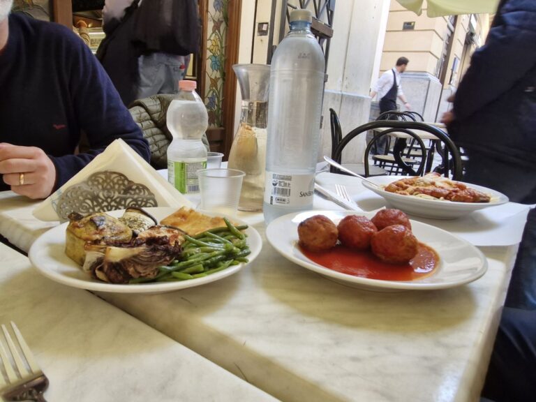 Where locals eat in Palermo. An outdoor restaurant table in Palermo featuring plates of traditional Sicilian food, including large meatballs in tomato sauce, a pasta dish, and a plate with green beans, grilled vegetables, and a slice of focaccia. Two bottles of water and a glass carafe sit on the marble table, with the entrance to Focacceria del Massimo visible in the background.