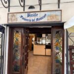 Gdzie zjeść w Palermo. Wejście do Basile Focacceria del Massimo w Palermo – otwarte drewniane drzwi z kolorowymi witrażami, nad nimi ceramiczny szyld z nazwą restauracji; w środku widoczna lada i fragment wnętrza.