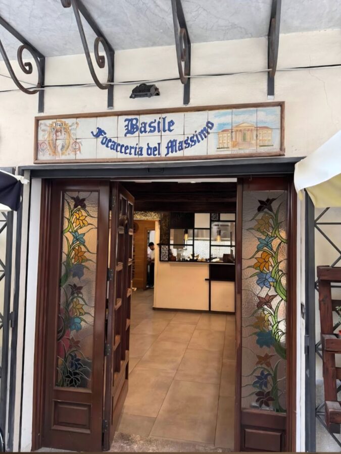 The entrance to Basile Focacceria del Massimo where locals eat in Palermo, featuring a traditional tiled sign above dark wood doors. The glass door panels are decorated with colorful stained-glass floral patterns, offering a view into the bright, tiled interior and the food service counter