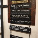 Handwritten Italian menu board hanging on a wall, listing pasta dishes and daily specials including ravioli, lasagne with pistachio pesto and mortadella, and anelletti al forno.