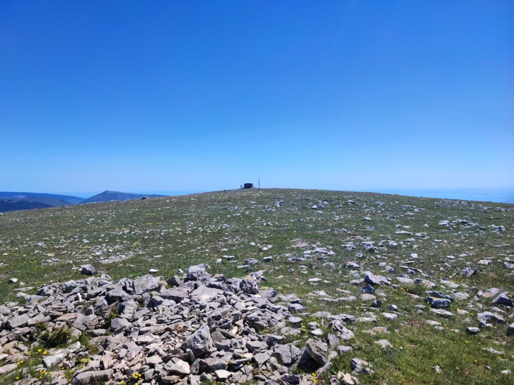 Monte Calvo Gargano, the highest peak in Gargano, featuring the stone refuge and cross against a clear blue sky. The foreground shows the karst landscape with scattered limestone rocks and wild herbs typical of the Puglia highlands.
