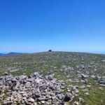 Monte Calvo Gargano, the highest peak in Gargano, featuring the stone refuge and cross against a clear blue sky. The foreground shows the karst landscape with scattered limestone rocks and wild herbs typical of the Puglia highlands.
