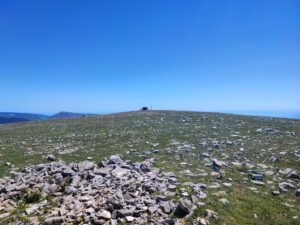 Monte Calvo Gargano, the highest peak in Gargano, featuring the stone refuge and cross against a clear blue sky. The foreground shows the karst landscape with scattered limestone rocks and wild herbs typical of the Puglia highlands.