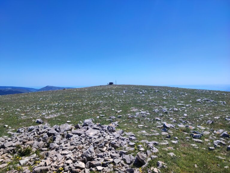 Monte Calvo Gargano, the highest peak in Gargano, featuring the stone refuge and cross against a clear blue sky. The foreground shows the karst landscape with scattered limestone rocks and wild herbs typical of the Puglia highlands.