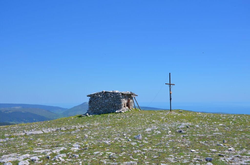 A small, primitive stone shelter built from dry limestone rocks sits on the flat, grassy summit of Monte Calvo. Next to the shelter stands a tall, thin metal cross, with the surrounding hills of Gargano and the blue sea visible in the distance under a clear sky.