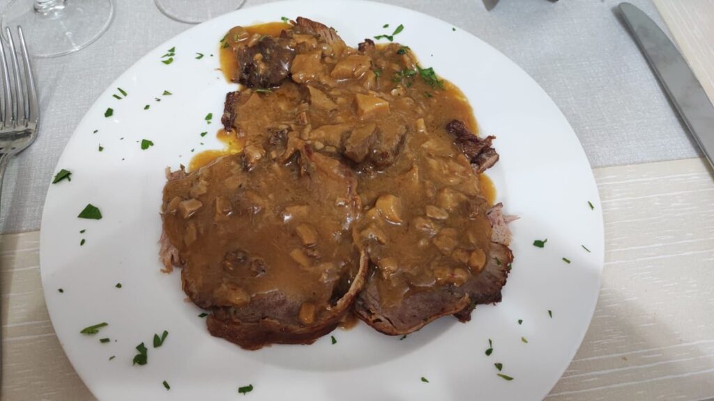 Slices of roast beef covered in a thick brown gravy with mushroom bits, served on a white plate and garnished with chopped parsley. The plate sits on a light-colored tablecloth next to a fork and knife.
