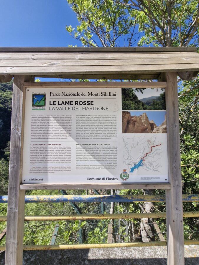 An information board at the start of the trail to Le Lame Rosse Marche hike, located near Lake Fiastra. The board features text in Italian and English, a map of the route, and photos of the local landscape and geological formations. It is mounted on a wooden frame outdoors, with green trees and a yellow metal railing visible in the background under a bright, sunny sky.
